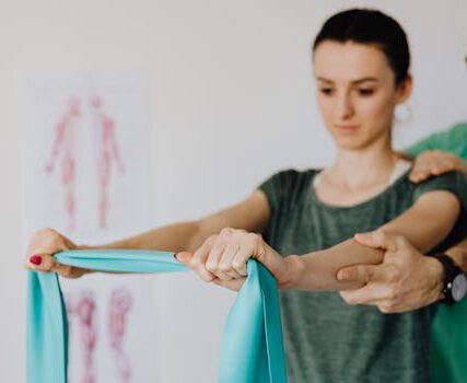 Leistungen Crop osteopath in uniform and eyeglasses supporting shoulder and reached arm of young woman with elastic tape in doctor office in hospital on blurred background